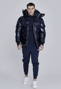 Námořnická puffer bunda s lesklým povrchem a kapucí, nošená přes odpovídající svetr a kalhoty. Bílé tenisky dotvářejí outfit.