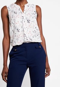 Ärmlös vit blus med röd och svart blommig tryck, kombinerad med högmidjade marinblå byxor med knappade fickor.
