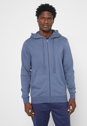 Man met een donkere huid die een lichtblauwe capuchontrui met rits en voorzakken en donkerblauwe broek draagt, staand voor een effen witte achtergrond.