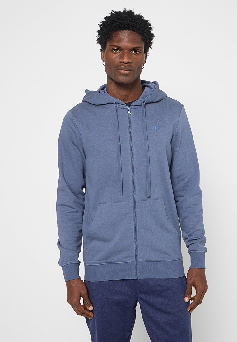 Homme à la peau foncée portant un sweat à capuche bleu clair zippé avec poches avant et un pantalon bleu marine, debout devant un fond blanc uni.