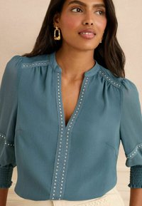 Blusa teal con scollo a V, maniche a puff e decorazioni di borchie argentate lungo la piastra. Tessuto leggero con finitura testurizzata.