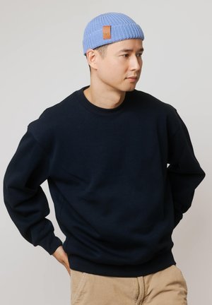 Mężczyzna noszący jasnoniebieską dzianinową czapkę i czarną bluzę, stojący z rękami na biodrach, patrzący w bok na jednolitym tle.