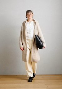 Manteau texturé beige clair avec capuche, chemise blanche à boutons, pantalon crème et sac en cuir noir. Le mannequin porte des chaussures noires, marchant devant un fond uni.