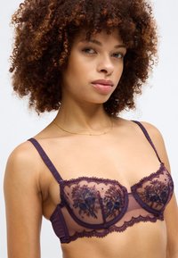 Purple lace bra with floral embroidery, sheer cups, scalloped edges, and adjustable straps. Textured fabric and delicate detailing visible.