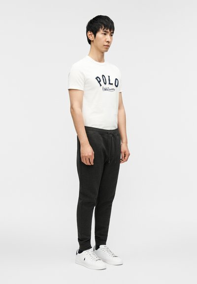 Λευκό μπλουζάκι με μπλε σκούρο λογότυπο "POLO", σκούρο γκρι φόρμα και λευκά αθλητικά παπούτσια με μαύρες λεπτομέρειες. Στάση όρθια, ουδέτερο φόντο.