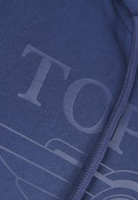 Navy sininen puuvillakangas, jossa on tekstuuripinta, suuria, hillitysti painettuja kirjaimia ja siipimäinen muotoiluelementti.