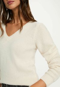 Maglione a V, di colore crema, realizzato in maglia con una texture morbida. Presenta maniche lunghe con dettagli in pizzo delicato ai polsini.