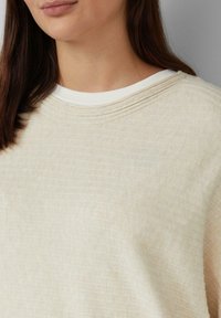 Jasno beżowy, teksturowany sweatshirt z gładką, białą okrągłą szyją i subtelnymi poziomymi liniami na tkaninie.