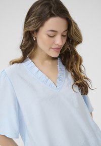 Haut bleu clair à manches courtes, avec un décolleté en V orné de volants, et fabriqué dans un tissu doux et légèrement texturé.