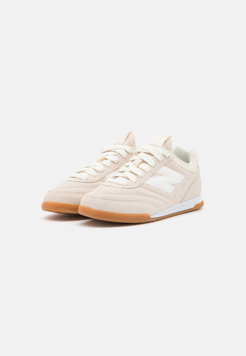 Baskets de sport beige clair avec un dessus en suède texturé, accent de logo blanc et semelle en caoutchouc couleur caramel. Présente des détails cousus et des lacets ronds.