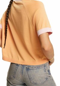 Kortærmet, cropped shirt i blødt ferskenfarvet bomuld, med lyserødt ribbet trim ved ærmerne og forneden. Sammen med lyseblå jeans.