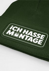 Dark green beanie made of ribbed fabric, featuring a prominent white patch with text "ICH HASSE MONTAGE" and a cartoon penguin design.