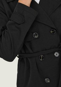 Trench-coat noir avec un design croisé à double boutonnage, de grands boutons noirs, des poches latérales et une ceinture à la taille pour une silhouette ajustée.