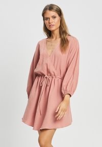 Šifonasta obleka roza barve, ki se ovija okoli telesa, iz lahkotnega materiala z dolgimi pufastimi rokavi, vrvico za povlečenje v pasu in obliko z V-izrezom, ki odlikuje tekoča silhueta.