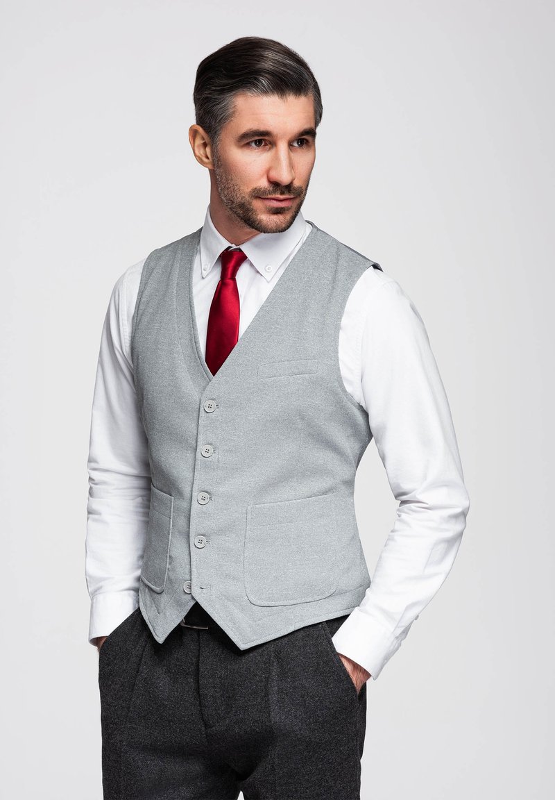 Homme aux cheveux foncés portant une chemise blanche, une cravate rouge, un gilet gris et un pantalon foncé, debout les mains dans les poches, regardant sur le côté.