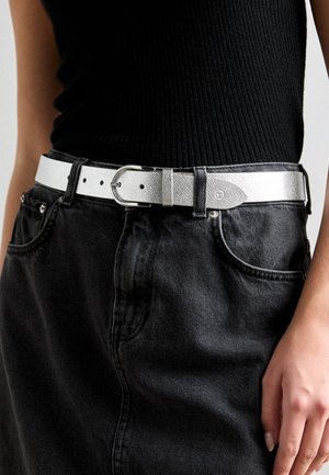 Ez egy ezüst, textúrált öv, téglalap alakú fém csattal, fekete farmer szoknya fölött viselve, öt övhurokkal és minimális részletezéssel.