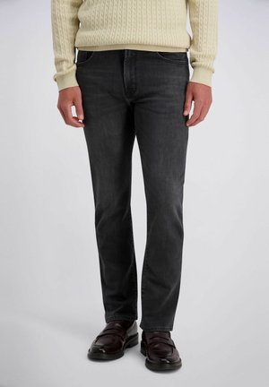 Mann trägt dunkelgraue Jeans, braune Ledermokassins und einen cremefarbenen Zopfpullover, steht vor einem schlichten Hintergrund.