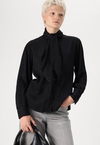 Svart knapp-up skjorta med vertikala ränder, knytsnodd, lång ärm. Matchas med ljusgrå jeans och en svart väska. Slät textur.