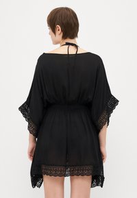 Cubierta negra con encaje, de corte holgado, mangas tipo kimono y un lazo en el cuello. Presenta material transparente y una cintura fruncida.