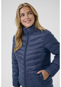 Jūros mėlynasis užsegamas pūkinis švarkas su aukštu apykakle, užtrauktuku ir šoninėmis kišenėmis. Lygus, blizgus audinys su prigludusiu siluetu.