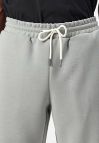 Lys grå sweatpants med elastisk talje, med en hvid snor der er bundet i en knude. Glat tekstur og tætsiddende form.