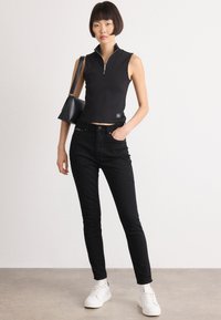 Černý bezrukávový crop top s předním zipem, zkombinovaný s vypasovanými černými džíny a bílými teniskami. Na rameni visí malá kabelka.