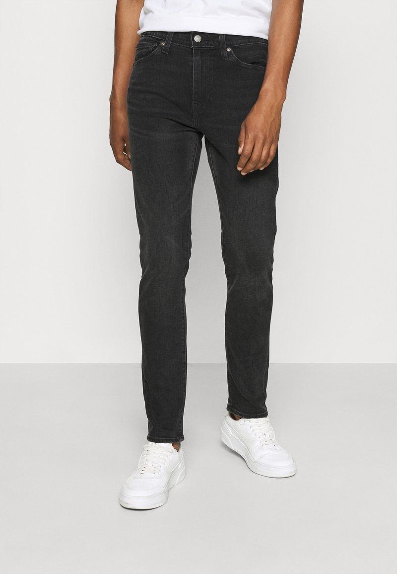 Man med åtsittande svarta jeans, vita sneakers och en vit t-shirt, stående mot en enfärgad ljus bakgrund.