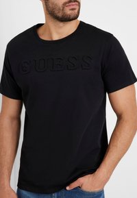 T-shirt in cotone nero con collo a giro e logo "GUESS" in rilievo sul davanti. Maniche corte e una texture liscia.