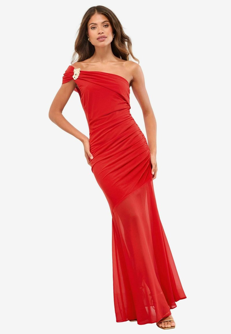 Czerwona sukienka na jedno ramię wykonana z elastycznego materiału, z ruchomymi detalami i przezroczystym wykończeniem na dole, ozdobiona dekoracyjną broszką.