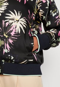 Zwarte bomberjack met een bloemenpatroon in roze, wit en groen. Heeft geribde marineblauwe manchetten en een lichtgroene accent langs de zoom.
