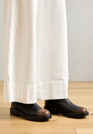 Mustad musta värvi ankle boots pruunide varvastega, valmistatud nahast, millel on ümar kuju ja lame kummitald. Kantud valgete laia jalaga pükstega.