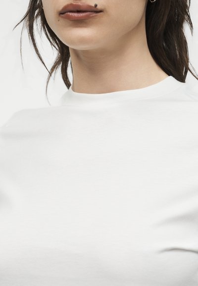 Κοντινή λήψη του κάτω μέρους προσώπου και του λαιμού μιας γυναίκας που φοράει λευκό πουκάμισο, με μια χαρακτηριστική ελιά πάνω από το άνω χείλος και ένα μικρό σκουλαρίκι κρίκο.