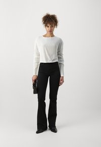 Un top long à manches longues blanc côtelé associé à un jean évasé noir. Le mannequin tient un petit sac à main noir et porte des chaussures noires à bouts pointus.