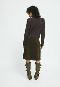 Brauner Kabelstrickpullover, olivfarbigem Cordrock und grünen, kniehohen Stiefeln mit Schnallenakzenten. Schlichte, strukturierte Stoffe in gedämpften Farbtönen.