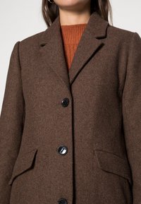 Frau in einem braunen Wollmantel mit schwarzen Knöpfen über einem rostfarbenen Strickpullover, der Oberkörper und Hals zeigt.