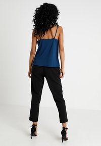 Donna con capelli ricci che indossa un top senza maniche blu, pantaloni neri arrotolati e scarpe con tacco nere, in piedi contro uno sfondo bianco uniforme.