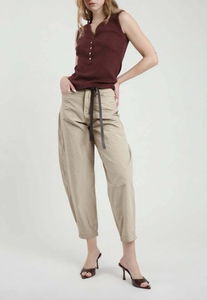 Donna che indossa un top marrone senza maniche, pantaloni beige a vita alta con cintura nera e sandali marroni con tacco alto, in piedi su sfondo bianco.