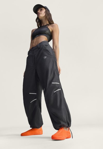 Žena u crnom crop topu, prostranim crnim hlačama s reflektirajućim detaljima, svijetlim narančastim slip-on tenisicama i crnom kapom, pozira protiv jednostavne pozadine.