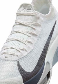 Nike sneaker featuring a white knit upper with textured accents, a black swoosh design, and a gray sole with visible cushioning.