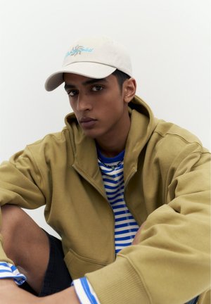 Persoană tânără purtând o șapcă bej, hanorac cu fermoar maro deschis și cămașă în dungi albastre și albe, așezată cu un genunchi ridicat pe fundal simplu.