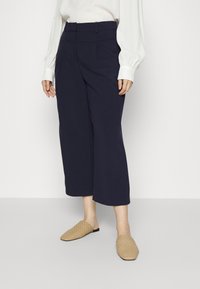 Mujer con pantalones cortos de pierna ancha en color marino, blusa blanca de manga larga y zapatos de lona beige de tipo slip-on, de pie sobre una superficie lisa.
