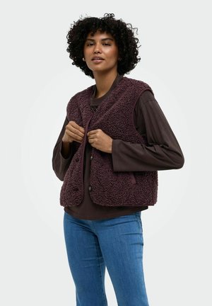 Frau mit lockigem Haar, die ein dunkelbraunes Langarmshirt, eine violette strukturierte Weste und blaue Jeans trägt und die Knöpfe der Weste zumacht.