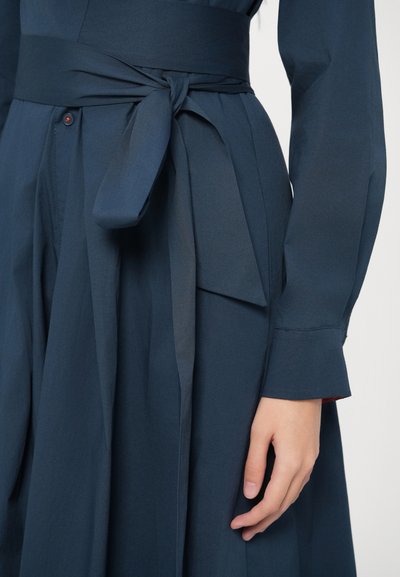 Granatowa sukienka z przewiązanym paskiem w talii, z długimi rękawami i gładką, teksturowaną tkaniną. Guziki z subtelnymi czerwonymi akcentami.