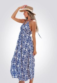 Vestido maxi de estampado azul y blanco con escote halter y tela fluida. Modelo usando un sombrero de paja, con la mano posicionada cerca de la cabeza.