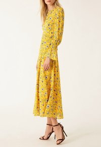 Femme portant une robe midi jaune à fleurs à manches longues et des sandales à talons hauts marron foncé, debout devant un fond uni.
