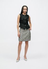 Schwarze Tweedweste mit zwei Fronttaschen und silbernen Knöpfen, kombiniert mit einem weißen und schwarzen Hahnentritt-Minirock, abgerundet durch hellfarbene High Heels.