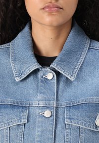 Jachetă din denim de culoare albastru deschis, cu un guler structurat, nasturi metalici argintii și două buzunare frontale cu detalii texturate subtile.