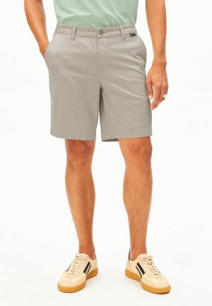 Uomo indossa pantaloncini casual grigio chiaro con tasche, abbinati a sneakers beige con strisce nere e suola color gomma, in piedi davanti a uno sfondo bianco.