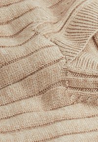 Tissu tricoté beige avec des motifs horizontaux côtelés et texturés, présentant des épaisseurs variables et une apparence douce et confortable.