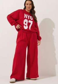Rode sweatshirt met witte print "NYC 97", gecombineerd met bijpassende rode broek met wijde pijpen. Zacht materiaal met een ontspannen pasvorm en een elastische taille. Witte schoenen.